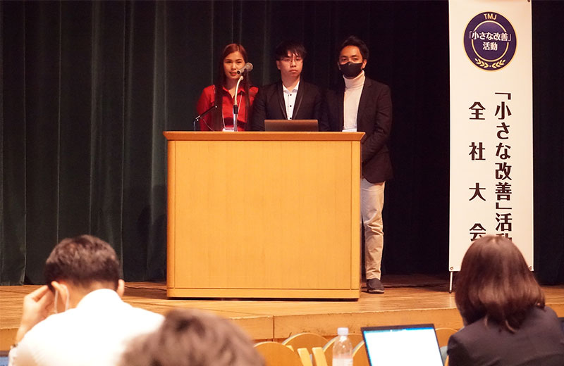 Three individuals at a podium, speaking into a microphone. Symbolizes a Kaizen event or conference.