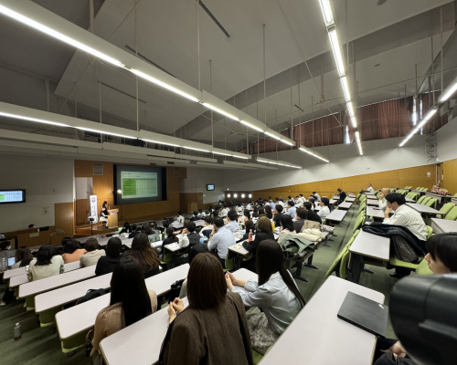 A large lecture hall filled with attendees from the BPO services industry, listening attentively to a speaker presenting at the podium during a Kaizen conference. The event focuses on continuous improvement practices within BPO services, with slides displayed on a large screen.