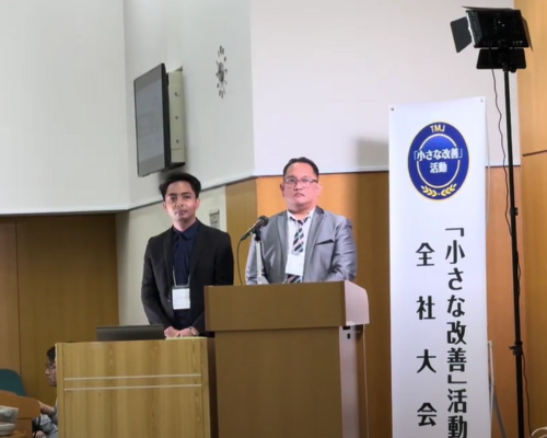 Two leaders from the BPO services sector stand behind a podium, delivering a presentation at a Kaizen event. A banner beside them highlights the focus on continuous improvement activities.