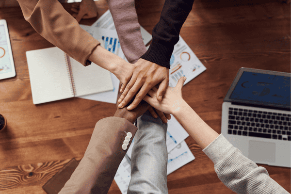 Hands of a diverse team stacked together above work documents and a laptop, symbolizing teamwork and trust, highlighting why TMJP Philippines is the ideal outsourcing partner.